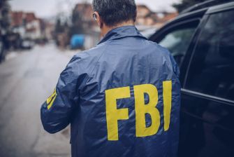 A person wearing a blue jacket with "FBI" printed in yellow on the back, standing on a street near a vehicle.