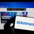 A person holding a smartphone displaying the Ticketmaster logo in front of a computer screen showcasing concert ticket promotions. A person holding a smartphone displaying the Ticketmaster logo in front of a computer screen showcasing concert ticket promotions.