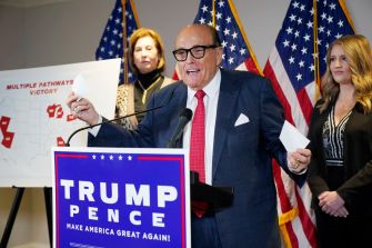 Rudy Giuliani speaking at a press conference with a "Trump Pence" backdrop, discussing election-related claims, while two women stand behind him.