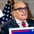 Rudy Giuliani speaking at a press conference, holding papers and surrounded by American flags. Rudy Giuliani speaking at a press conference, holding papers and surrounded by American flags.
