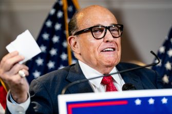 Rudy Giuliani speaking at a press conference, holding papers and surrounded by American flags.