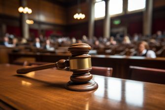 A wooden gavel resting on a desk in a courtroom with blurred figures of people in the background.