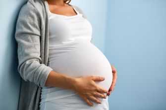 A pregnant woman in a white dress is standing with her hands on her belly against a blue wall.