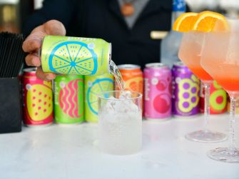 A hand pouring Poppi prebiotic soda into a glass, with various colorful cans of Poppi in the background.