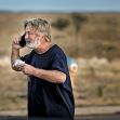 A man speaking on a mobile phone outdoors, wearing a dark shirt, with a blurred landscape in the background. A man speaking on a mobile phone outdoors, wearing a dark shirt, with a blurred landscape in the background.