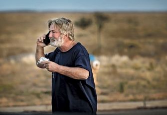 A man speaking on a mobile phone outdoors, wearing a dark shirt, with a blurred landscape in the background.
