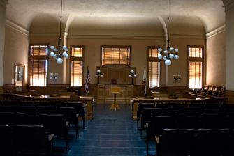 A view of a courtroom with empty benches, wooden furnishings, and windows allowing natural light.