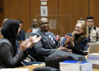 A man sitting in a courtroom surrounded by supporters, smiling and clapping as he celebrates his exoneration after being wrongfully convicted.
