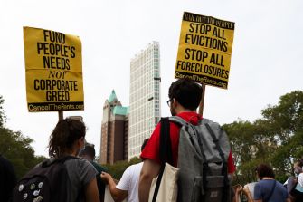 Protesters holding signs advocating for rent relief and a halt to evictions, with a city skyline in the background.