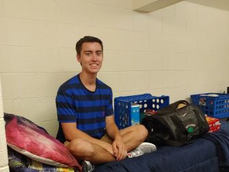 A young male student sitting cross-legged on a bed in a dorm room, smiling at the camera.