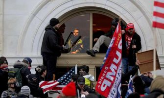 A group of individuals, some holding flags, breach the Capitol building during the January 6 riot, while others look on from inside.