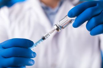 A person in a lab coat holding a vaccination vial and syringe, wearing blue gloves.