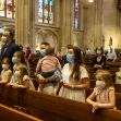 A group of masked individuals of various ages stands in a church pew, observing their surroundings. Stained glass windows are visible in the background. A group of masked individuals of various ages stands in a church pew, observing their surroundings. Stained glass windows are visible in the background.