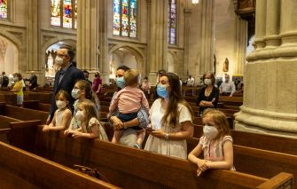 A group of masked individuals of various ages stands in a church pew, observing their surroundings. Stained glass windows are visible in the background.