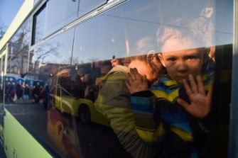 A child looking out from a bus window, with reflections of people and a queue visible outside, amidst the ongoing Ukrainian refugee crisis.