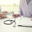 A person handing cash to a healthcare provider while another writes on a clipboard, with a stethoscope nearby, symbolizing healthcare transactions. A person handing cash to a healthcare provider while another writes on a clipboard, with a stethoscope nearby, symbolizing healthcare transactions.