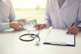 A person handing cash to a healthcare provider while another writes on a clipboard, with a stethoscope nearby, symbolizing healthcare transactions.