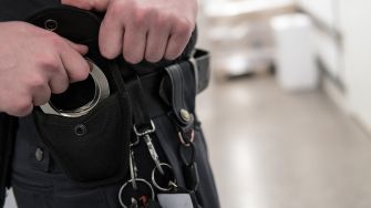 A close-up image of a prison guard's hands adjusting their handcuffs and utility belt.