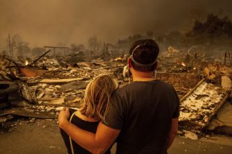 A couple stands together amidst the rubble of destroyed homes, surveying the devastation caused by a wildfire.