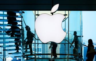 Apple logo displayed in a store with silhouettes of people walking in the background.