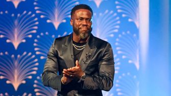Kevin Hart clapping during a performance, wearing a dark suit against a decorative background.
