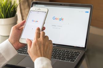 A person using a smartphone with the Google homepage displayed on a laptop in the background.