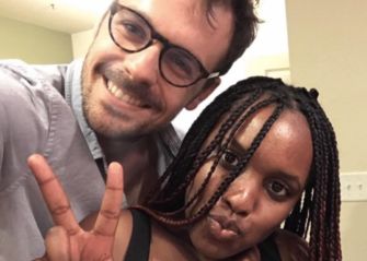 A man and a woman posing together for a selfie, both smiling, with the woman making a peace sign gesture.