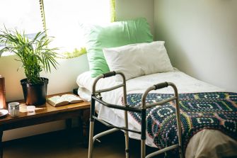 A nursing home's interior room featuring a bed with colorful blankets, a walker, a potted plant, and a small table with items, highlighting the environment where elderly care takes place.