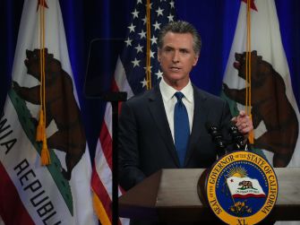 California Governor speaking at a podium with California state flags in the background.