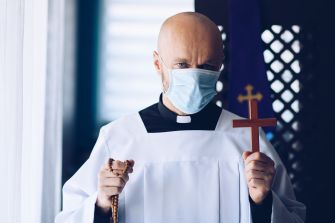 A masked priest holds a wooden cross and a rosary, symbolizing faith during the COVID-19 pandemic.