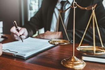 A close-up view of a lawyer's hands writing on legal documents with a scale of justice in the foreground.
