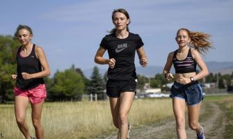 Three young female athletes running on a grassy path.
