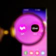 Logos of Lyft and Uber displayed on a smartphone screen, with a magnifying glass focusing on them. Logos of Lyft and Uber displayed on a smartphone screen, with a magnifying glass focusing on them.