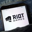 A smartphone displaying the Riot Games logo, partially hidden in a denim pocket. A smartphone displaying the Riot Games logo, partially hidden in a denim pocket.