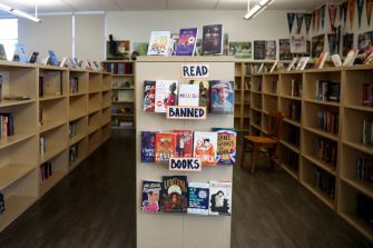 A bookshelf displaying various books with a sign that reads "READ BANNED BOOKS" in a library setting.