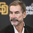 A man in a suit speaking at a podium in front of a logo for the San Diego Padres baseball team. A man in a suit speaking at a podium in front of a logo for the San Diego Padres baseball team.