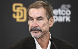 A man in a suit speaking at a podium in front of a logo for the San Diego Padres baseball team.