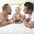 A same-sex couple playing with a smiling baby on a bed. A same-sex couple playing with a smiling baby on a bed.