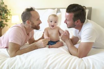 A same-sex couple playing with a smiling baby on a bed.
