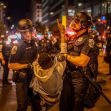 A police officer restrains a protester during a nighttime demonstration, with multiple police officers and emergency vehicle lights in the background. A police officer restrains a protester during a nighttime demonstration, with multiple police officers and emergency vehicle lights in the background.