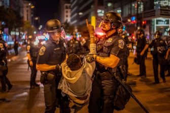A police officer restrains a protester during a nighttime demonstration, with multiple police officers and emergency vehicle lights in the background.