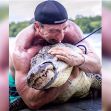 A muscular man wearing a cap is wrestling with a large alligator while holding it against his chest. A muscular man wearing a cap is wrestling with a large alligator while holding it against his chest.