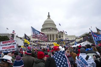 Crowd gathered at the U.S. Capitol during the January 6th riot, with flags and signs visible.