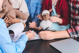Hands writing on a document at a table with children holding stuffed animals nearby.