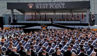 A large gathering of West Point cadets listening to a speaker at a ceremony, with the West Point banner displayed prominently in the background.