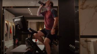 A man drinking from a water bottle while exercising on a Peloton bike in an upscale home setting.