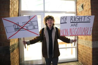 A student holding two signs outside a school, one crossed out with a building drawing and the other stating "MY RIGHTS ARE NON-NEGOTIABLE."