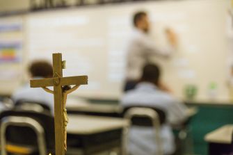 A wooden crucifix in the foreground with an out-of-focus teacher writing on a whiteboard in a classroom setting.