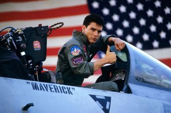 A pilot giving a thumbs-up in the cockpit of a fighter jet with an American flag in the background.