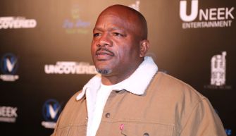 A man in a beige jacket stands in front of a black backdrop with the logos of Uneek Entertainment and the text "UNVEILED."